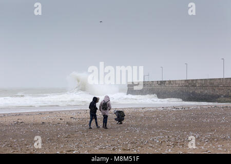 Ramsgate, Kent, UK 18th March, 2018. Three young people on the beach during a snow storm with a large wave behind them close to the harbour arm. Sue Holness Credit: Sue Holness/Alamy Live News Stock Photo