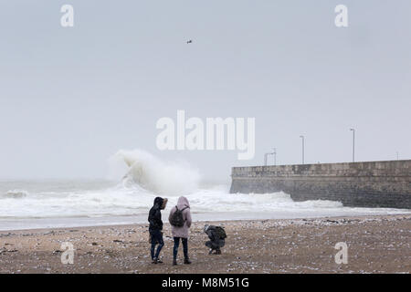 Ramsgate, Kent, UK 18th March, 2018. Three young people on the beach during a snow storm with a large wave behind them close to the harbour arm. Sue Holness Credit: Sue Holness/Alamy Live News Stock Photo