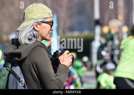 Senior woman wearing a ball cap holding a camera in her hand and smiling.  She is watching the Bellingham, Washington St. Patrick's Day Parade on Marc Stock Photo