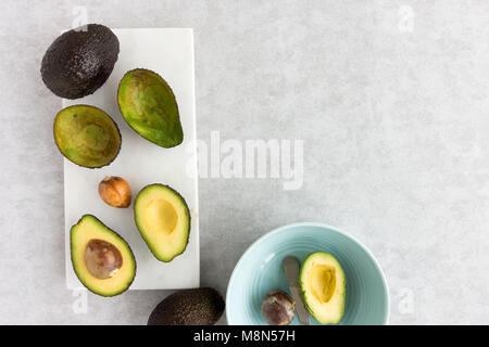 Avocados, whole, halved and scooped out with avocado tool on marble chopping board and in a blue bowl. Top View and copy space. Stock Photo