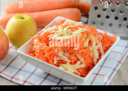 Concept of preparing a healthy salad - grated carrot with apple in a white platter on a background of graters, carrots and apples Stock Photo