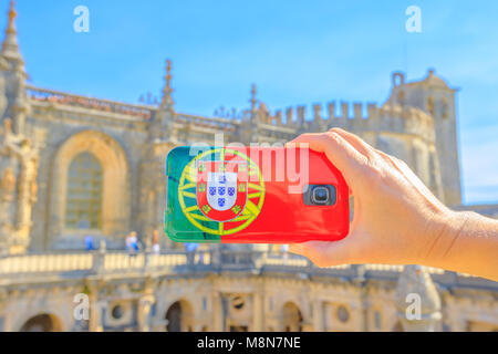 Tourism and travel concept in Portugal. Close up of mobile phone with Portugal flag cover taking photos of tourist place in Templar city. Convent of Christ or Templar fortress on blurred background. Stock Photo