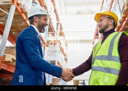 Waist up portrait of handsome mature businessman shaking hands with worker wearing hardhat standing against warehouse shelves in background Stock Photo