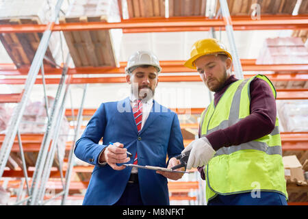 Waist up portrait of mature businessman talking to warehouse worker, discussing inventory on clipboard Stock Photo