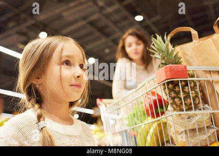 Warm-toned portrait of family doing grocery shopping in supermarket, focus on little girl standing by cart, copy space Stock Photo