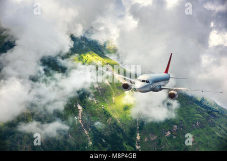 Aerial view of aircraft. Airplane is flying in clouds over mountains with forest at sunset. Landscape with passenger airplane, cloudy sky, trees. Pass