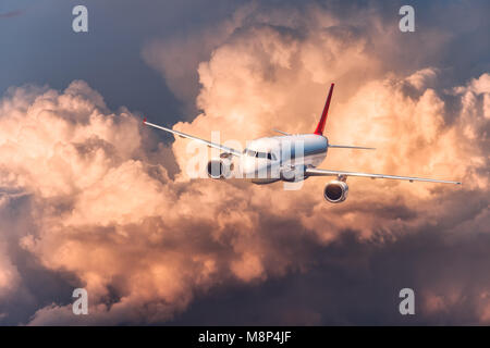 Beautiful airplane is flying in low clouds at colorful sunset. Scene with white passenger airplane sky with orange and red clouds. Passenger aircraft. Stock Photo