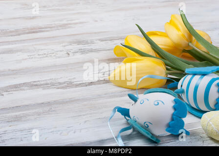 bouquet of yellow tulips and toys chikken easter eggs with a blue ribbon on vintage white wooden table background.  holidays concept. Copy space. Stock Photo
