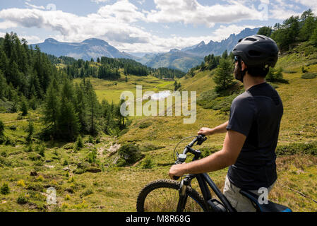 Young adult active man on mountain wearing bike helmet looking at scenic panorama holding electric bike in sunny summer day outdoor. Stock Photo
