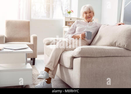 Charming elderly lady relaxing with coffee cup on couch Stock Photo