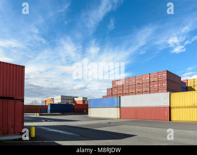 Dry cargo containers of different colors stacked in a shipping yard under a blue sky with white clouds. Stock Photo