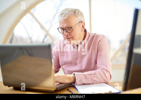 Retired man wearing shirt and sitting at desk while working on laptop from home. Stock Photo