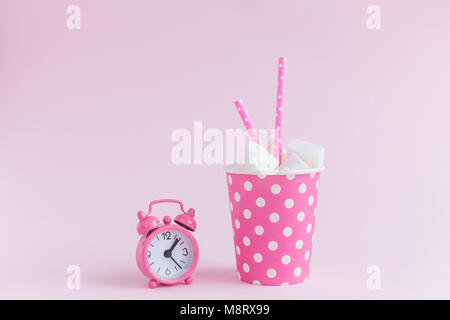Paper cup polka design full of sugar cubes with drinking straws and small alarm clock on pastel pink background minimal concept. Stock Photo