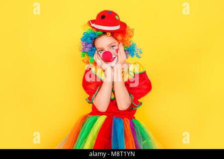 Funny little kid wearing clown costume touches cheeks and smiling. Studio shot, isolated on yellow background Stock Photo