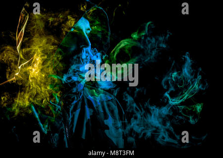A young man in a black hood smokes and breathes out the colored yellow and green  smoke from the vape on a black  isolated background Stock Photo