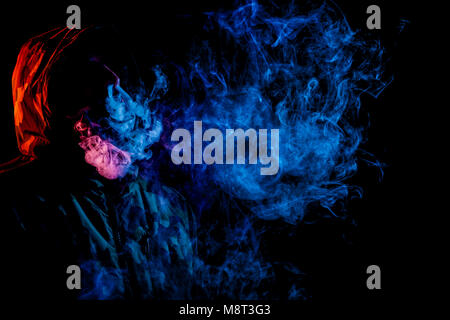 A young man in a black hood smokes and breathes out the colored blue and red smoke from the vape on a black isolated background Stock Photo