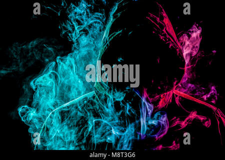 A young man in a black hood smokes and breathes out the colored  green  and pink smoke from the vape on a black isolated background Stock Photo