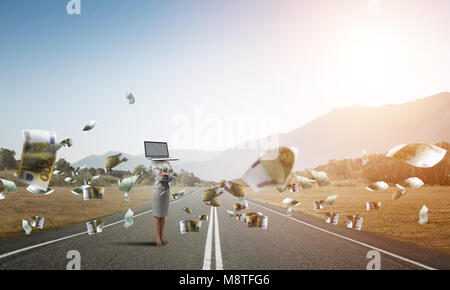 Business woman in suit with laptop instead of head keeping arms crossed while standing on the road among flying euro banknotes with beautiful landscap Stock Photo