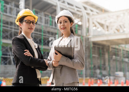 Two a professional female engineer looking at the camera with a confident smile. They both wearing yellow hard cap standing aside the building site. Stock Photo