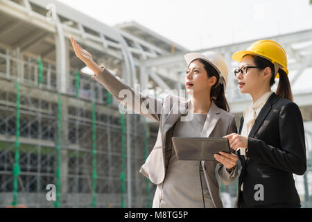 Female architect wearing white hard hat point showing the building project to the woman wearing a yellow hard hat. They both wearing suits standing as Stock Photo