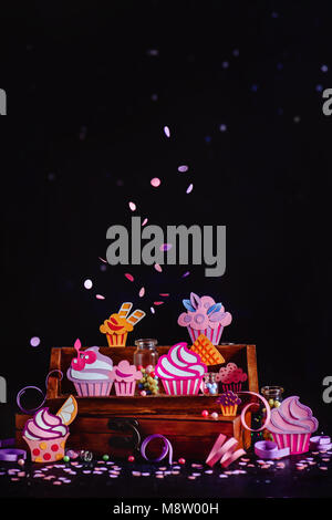 Celebration concept. Cupcakes collection on a wooden shelf with flying confetti and sprinkles. Colorful papercraft dessert assortment a dark backgroun Stock Photo