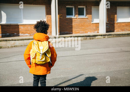 Little school boy in orange coat walking at the entrance of the school with a yellow backpack. Back view