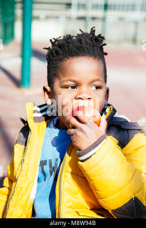 Cute little boy with a yellow coat eating a red apple at the school in a sunny day Stock Photo