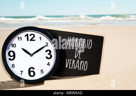 closeup of a clock and a signboard with the text horario de verano, summer time written in spanish, on the sand of a beach Stock Photo