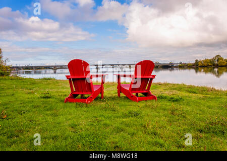 two empty red chaise longues stand on the grass opposite the river and the bridge Stock Photo