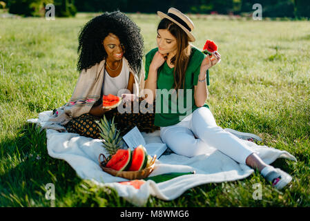 Outdoor portrait of two cheerful smiling pretty multi-race girl friends on the picnic. The african girl is showing to her charming friend something on the mobile while eating the watermelon. Stock Photo