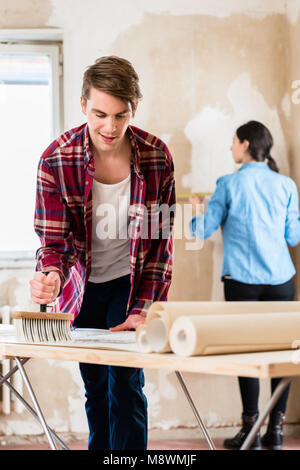 Young man applying adhesive to wallpaper while girlfriend is measuring the wall Stock Photo