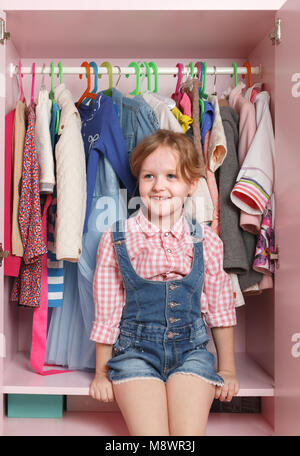 A little girl is sitting in a closet with a children's department. Storage system for children's things Stock Photo