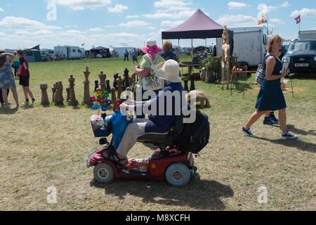 Woman in a mobility scooter driving past a big wooden chess set by a wood carver at the Gloucestershire Vintage Country Show in Cirencester 2016 Stock Photo