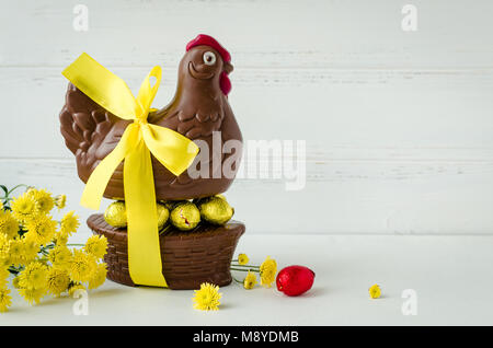 Easter composition with chocolate chicken in the nest and eggs with yellow ribbon and flowers on white shabby chic wooden background with place for te Stock Photo
