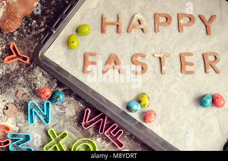 Easter baking concept: raw dough for biscuit, colorful eggs, flour, cutters in the shape of letters on a dark wooden table. Easter baking preparation. Stock Photo