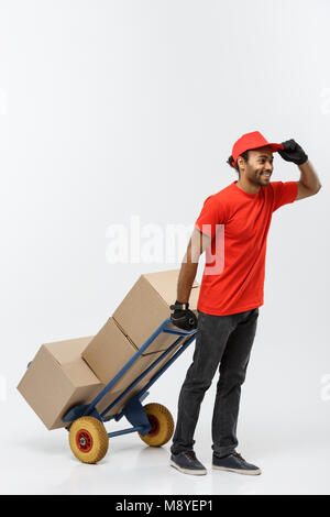 Delivery Concept - Portrait of Handsome African American delivery man or courier pushing hand truck with stack of boxes. Isolated on Grey studio Background. Copy Space. Stock Photo