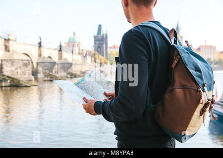A young tourist man with a backpack standing next to the river Vltava in Prague looks at the map and admires the architecture of the city. Charles Bridge is near. Travel around the Czech Republic. Stock Photo