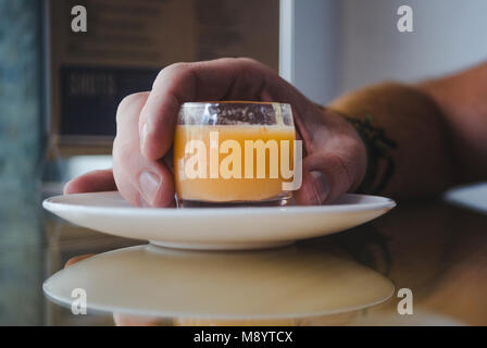 Young millennial man's hand holds healthy shot of functional organic fruit, vegetable and spice in a small glass on a white saucer in a health bar Stock Photo