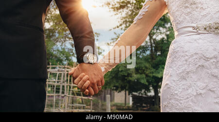 Close up of bride in white dress and groom in suit holding each others hands outdoors. Couple holding hands on their wedding day. Cropped shot with fo Stock Photo