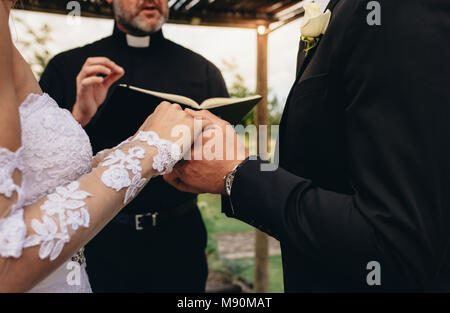 Close up of bride and groom holding each other's hands during outdoor wedding ceremony. Priest performing a wedding ceremony for couple outdoors. Stock Photo