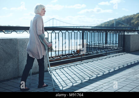 Worried senior woman staring somewhere while walking with crutches Stock Photo
