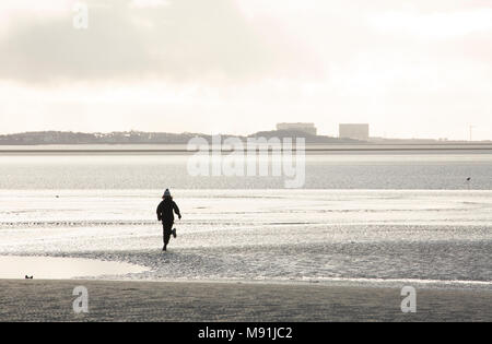 A youth running across sands at low tide at Jenny Brown’s Point near Silverdale UK with Heysham Nuclear Power station in the distance. January 2018 UK Stock Photo