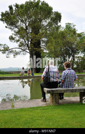 Romantic old couple sitting on park bench by lake. In front of them there is a couple of young lovers who fight. Stock Photo