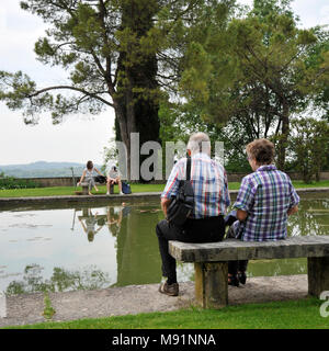 Romantic old couple sitting on park bench by lake. In front of them there is a couple of young lovers who fight. Stock Photo