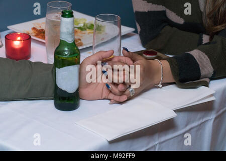 Young couple in love on a date hold hands across table after dinner. Empty plate of food, candle, drink glasses and generic beer bottle with white lab Stock Photo
