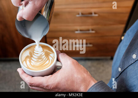 Professional barista pouring steamed milk from stainless steel tumbler into coffee cup making latte art on cappuccino Stock Photo