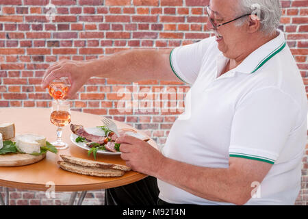 fat man eating a lot of unhealthy foods lots of cheese, meat and sausage products drinking glass of wine outdoor over a red brick wall Stock Photo