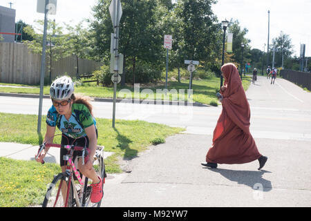 A difference in cultures, a Muslim woman in her hijab crossing the Midtown Greenway trail behind a woman on her bicycle. Minneapolis Minnesota MN USA Stock Photo