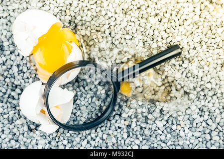 Broken egg with magnifying glass on gray gravel, asphalt. Risk of loss, ruin, investigation of the incident. Stock Photo