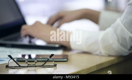 Closeup of eyeglasses on office table, female hands typing on laptop, eyestrain Stock Photo